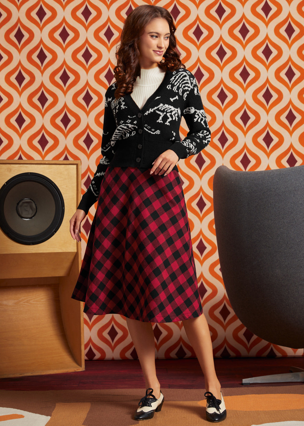 A woman models a black cardigan with white skeleton motifs and a red and black plaid midi skirt, standing in front of a retro orange and brown patterned wallpaper background.