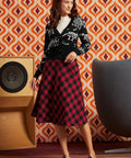 A woman models a black cardigan with white skeleton motifs and a red and black plaid midi skirt, standing in front of a retro orange and brown patterned wallpaper background.