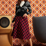 A woman models a black cardigan with white skeleton motifs and a red and black plaid midi skirt, standing in front of a retro orange and brown patterned wallpaper background.