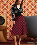 A woman models a black cardigan with white dinosaur bones and a red and black plaid midi skirt, paired with black and white Mary Jane shoes, set against a retro orange and brown patterned wallpaper background.