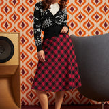 A woman models a black cardigan with white dinosaur bones and a red and black plaid midi skirt, paired with black and white Mary Jane shoes, set against a retro orange and brown patterned wallpaper background.