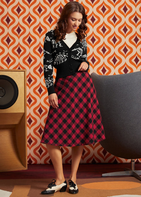 A woman models a black cardigan with white dinosaur bones and a red and black plaid midi skirt, paired with black and white Mary Jane shoes, set against a retro orange and brown patterned wallpaper background.