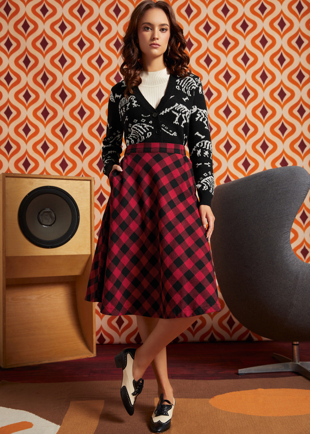 Woman wearing red and black buffalo check midi skirt with black dinosaur cardigan and black-and-white oxford shoes, front view with hand in side pocket