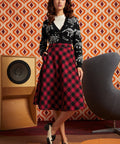 Woman wearing red and black buffalo check midi skirt with black dinosaur cardigan and black-and-white oxford shoes, front view with hand in side pocket