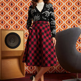 Woman wearing red and black buffalo check midi skirt with black dinosaur cardigan and black-and-white oxford shoes, front view with hand in side pocket