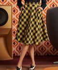 Woman wearing mustard and black check midi skirt with black lace blouse and black-and-white oxford shoes, front view with hands in side pockets