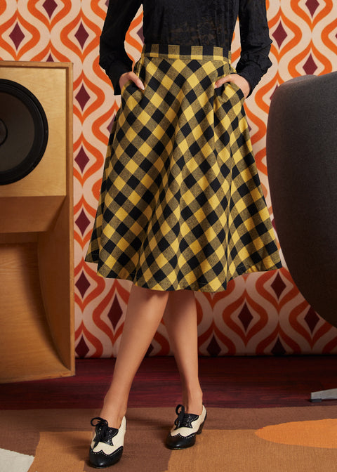 Woman wearing mustard and black check midi skirt with black lace blouse and black-and-white oxford shoes, front view with hands in side pockets