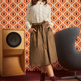 A woman stands in a retro-styled room wearing a cream textured knit top, a brown suede-like tie-waist midi skirt, and dark burgundy lace-up ankle boots.