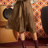A woman wearing a brown midi skirt with a matching waist tie, paired with a cream patterned top and burgundy lace-up ankle boots, standing in front of a retro orange and white patterned wallpaper background.
