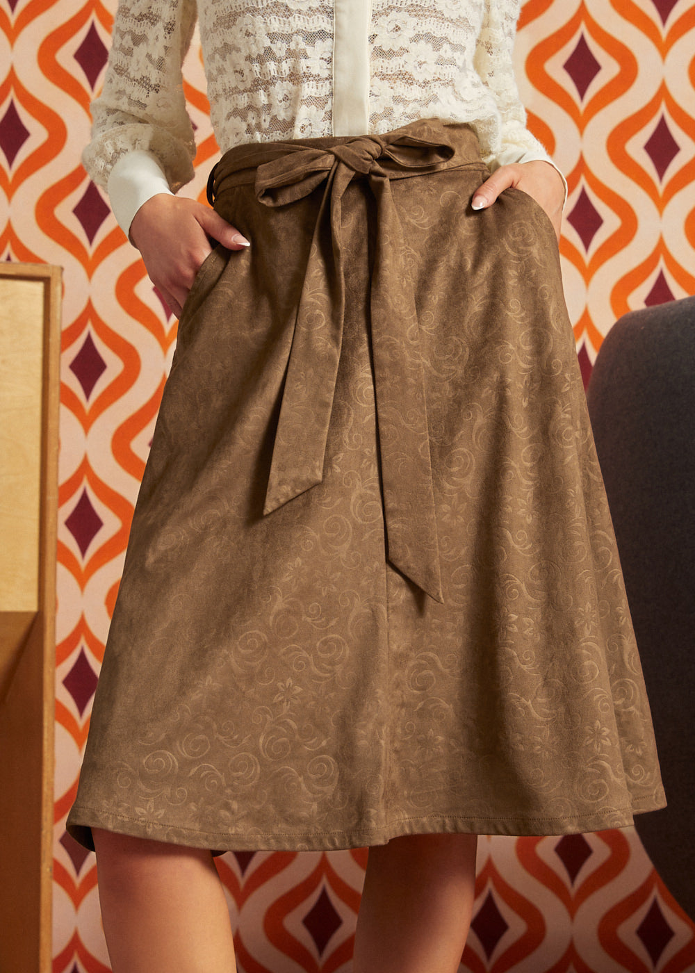 Woman wearing a mocha brown floral A-line skirt with a tied waist sash and side pockets, styled with an ivory lace blouse against an orange retro wallpaper background.