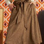 Woman wearing a mocha brown floral A-line skirt with a tied waist sash and side pockets, styled with an ivory lace blouse against an orange retro wallpaper background.