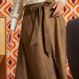 A brown midi skirt with a subtle jacquard pattern and a matching self-tie belt, worn by a model in front of a retro orange and white patterned wallpaper background.