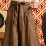 A brown textured A-line skirt with a matching fabric tie belt at the waist, worn by a model in front of a retro orange and white patterned wallpaper background.