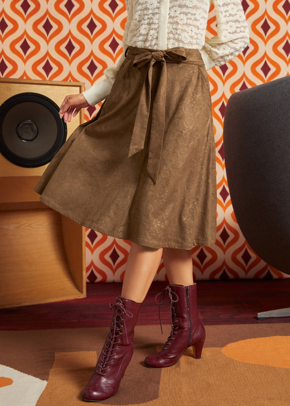 A woman wearing a brown textured midi skirt with a tie waist, a cream knit top, and burgundy lace-up ankle boots, standing in a retro-styled room with patterned wallpaper and a speaker.