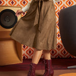 A woman wearing a brown textured midi skirt with a tie waist, a cream knit top, and burgundy lace-up ankle boots, standing in a retro-styled room with patterned wallpaper and a speaker.