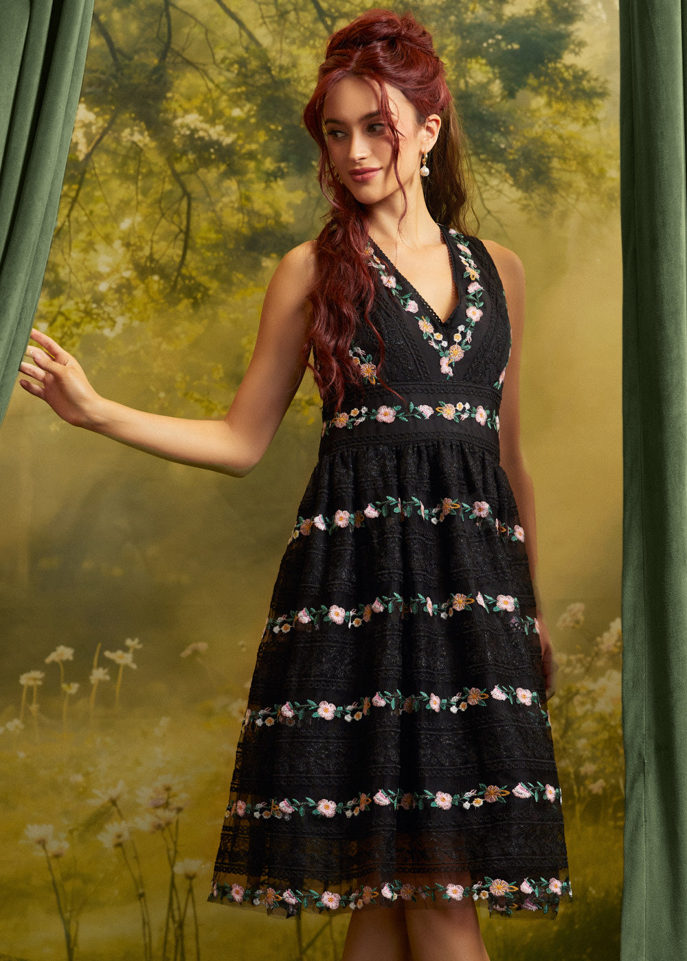 A woman wearing a sleeveless black midi dress with a V-neckline and horizontal floral embroidered bands, posed in front of a green forest backdrop with curtains.