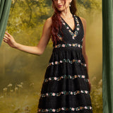 A woman wearing a sleeveless black midi dress with a V-neckline and horizontal floral embroidered bands, posed in front of a green forest backdrop with curtains.