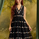 A woman modeling a sleeveless black ModCloth midi dress with a V-neckline and horizontal floral lace bands, posed against a soft-focus green and gold forest backdrop.