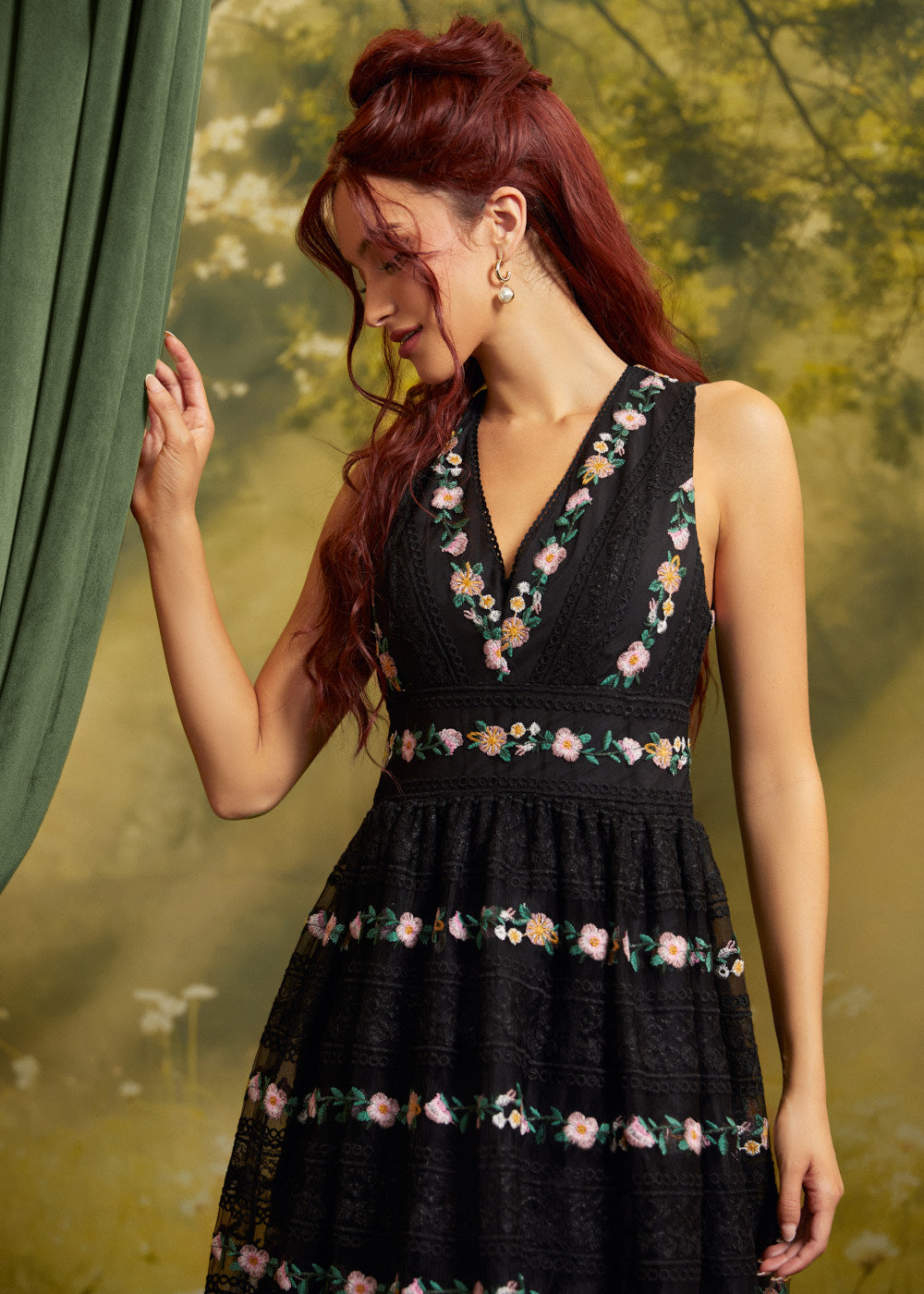 A woman with red hair wearing a sleeveless black dress with colorful floral embroidery, standing next to a green curtain against a soft-focus green background.