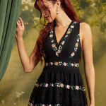 A woman with red hair wearing a sleeveless black dress with colorful floral embroidery, standing next to a green curtain against a soft-focus green background.