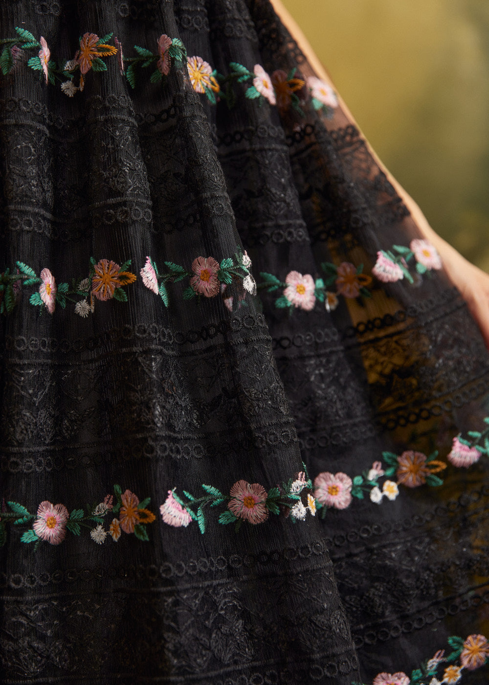 Close-up of a sheer black lace skirt showing horizontal floral embroidery, intricate lace patterns, and pink, yellow, and green botanical details.