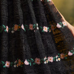Close-up of a sheer black lace skirt showing horizontal floral embroidery, intricate lace patterns, and pink, yellow, and green botanical details.