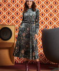 A woman models a long-sleeve midi dress with a black, gray, and beige paisley pattern, styled with a pink headband and burgundy lace-up boots, set against a retro orange and brown patterned wallpaper background.