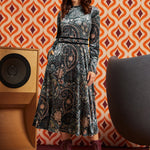 A woman models a long-sleeve midi dress with a black, gray, and beige paisley pattern, styled with a pink headband and burgundy lace-up boots, set against a retro orange and brown patterned wallpaper background.