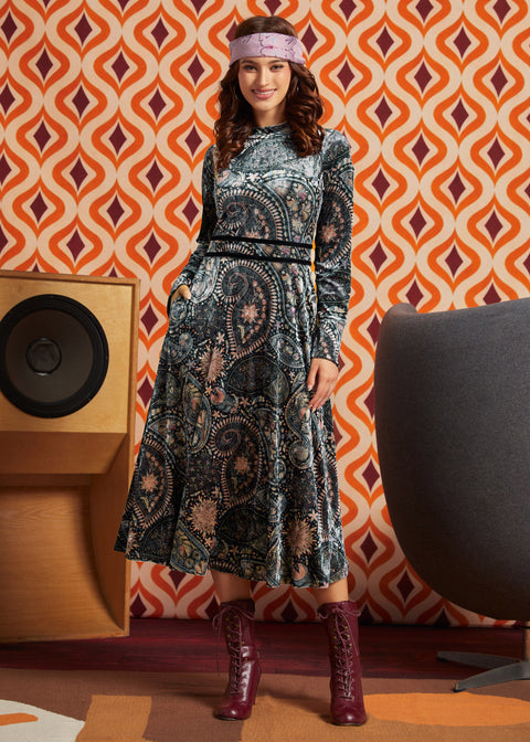 A woman models a long-sleeve midi dress with a black, gray, and beige paisley pattern, styled with a pink headband and burgundy lace-up boots, set against a retro orange and brown patterned wallpaper background.