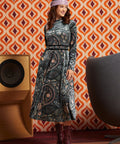 A woman stands in a retro-styled room wearing a long-sleeve midi dress with a dark paisley pattern and burgundy lace-up ankle boots. The background features orange and cream geometric wallpaper, a wooden speaker, and a gray chair.