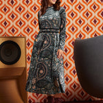 A woman stands in a retro-styled room wearing a long-sleeve midi dress with a dark paisley pattern and burgundy lace-up ankle boots. The background features orange and cream geometric wallpaper, a wooden speaker, and a gray chair.