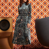 A woman stands in a retro-styled room wearing a long-sleeve midi dress with a dark paisley pattern and burgundy lace-up ankle boots. The background features orange and cream geometric wallpaper, a wooden speaker, and a gray chair.