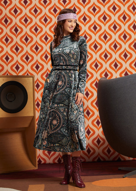 A woman stands in a retro-styled room wearing a long-sleeve midi dress with a dark paisley pattern and burgundy lace-up ankle boots. The background features orange and cream geometric wallpaper, a wooden speaker, and a gray chair.
