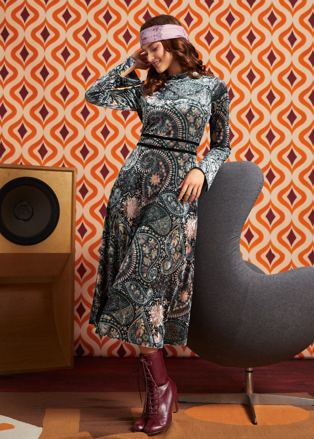 A woman models a long-sleeve midi dress with a complex paisley and floral pattern in blue, gray, and pink, paired with a pink headband and burgundy lace-up ankle boots. She is posed next to a modern gray chair in front of a retro orange and brown patterned wallpaper background.