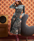 A woman models a long-sleeve midi dress with a complex paisley and floral pattern in blue, gray, and pink, paired with a pink headband and burgundy lace-up ankle boots. She is posed next to a modern gray chair in front of a retro orange and brown patterned wallpaper background.