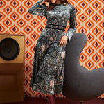A woman models a long-sleeve midi dress with a complex paisley and floral pattern in blue, gray, and pink, paired with a pink headband and burgundy lace-up ankle boots. She is posed next to a modern gray chair in front of a retro orange and brown patterned wallpaper background.