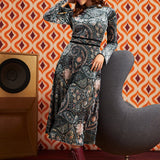 A woman models a long-sleeve midi dress with a complex paisley and floral pattern in blue, gray, and pink, paired with a pink headband and burgundy lace-up ankle boots. She is posed next to a modern gray chair in front of a retro orange and brown patterned wallpaper background.