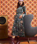 A woman models a long-sleeved midi dress with a dark blue and gray paisley pattern, styled with a pink headband and burgundy lace-up ankle boots, set against a retro orange and white patterned wallpaper background.