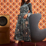 A woman models a long-sleeved midi dress with a dark blue and gray paisley pattern, styled with a pink headband and burgundy lace-up ankle boots, set against a retro orange and white patterned wallpaper background.