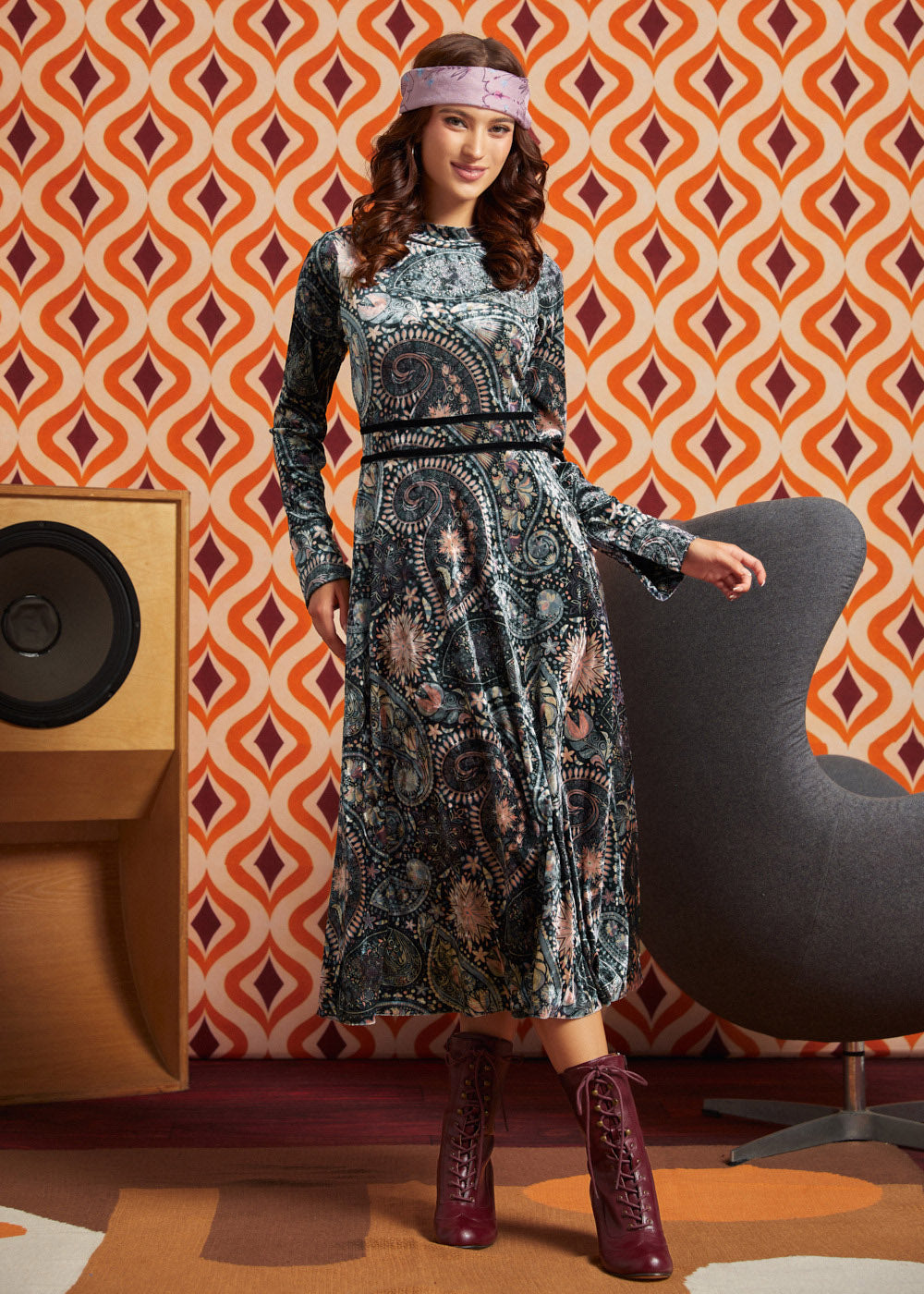A woman models a long-sleeve midi dress with a dark paisley pattern in black, blue, and beige tones, styled with a pink headband and burgundy lace-up boots, set against a retro orange and white patterned wallpaper background.