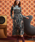 A woman models a long-sleeve midi dress with a dark paisley pattern in black, blue, and beige tones, styled with a pink headband and burgundy lace-up boots, set against a retro orange and white patterned wallpaper background.