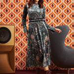 A woman models a long-sleeve midi dress with a dark paisley pattern in black, blue, and beige tones, styled with a pink headband and burgundy lace-up boots, set against a retro orange and white patterned wallpaper background.