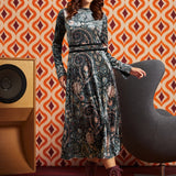 A woman models a long-sleeve midi dress with a dark paisley pattern in black, blue, and beige tones, styled with a pink headband and burgundy lace-up boots, set against a retro orange and white patterned wallpaper background.