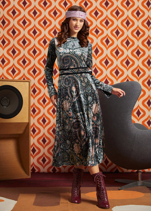 A woman models a long-sleeve midi dress with a dark paisley pattern in black, blue, and beige tones, styled with a pink headband and burgundy lace-up boots, set against a retro orange and white patterned wallpaper background.