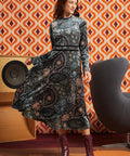 A woman modeling a long-sleeve ModCloth midi dress with an intricate paisley and floral pattern in black, gray, and muted blue tones, paired with dark red lace-up ankle boots, set against a retro orange and brown patterned wallpaper background.