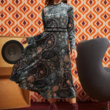 A woman modeling a long-sleeve ModCloth midi dress with an intricate paisley and floral pattern in black, gray, and muted blue tones, paired with dark red lace-up ankle boots, set against a retro orange and brown patterned wallpaper background.