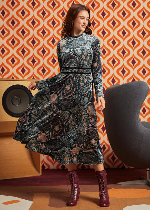 A woman modeling a long-sleeve ModCloth midi dress with an intricate paisley and floral pattern in black, gray, and muted blue tones, paired with dark red lace-up ankle boots, set against a retro orange and brown patterned wallpaper background.