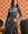Woman wearing dark paisley floral midi dress with long flared sleeves and black waist trim, front view with one hand in side pocket