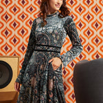 Woman wearing dark paisley floral midi dress with long flared sleeves and black waist trim, front view with one hand in side pocket