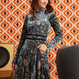 Woman wearing dark paisley floral midi dress with long flared sleeves and black waist trim, front view with one hand in side pocket